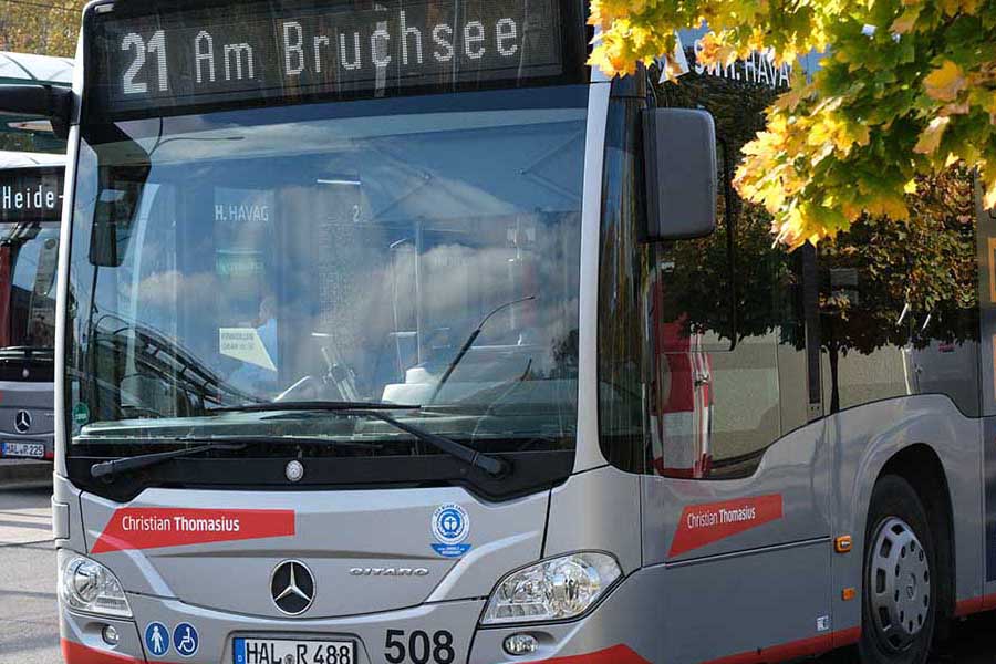 Nahaufnahme eines silbernen Mercedes-Stadtbusses mit rotem Akzent, Liniennummer 21 und Zielanzeige '21 Am Bruchsee', geparkt unter einem Baum mit herbstlich gefärbtem Laub
