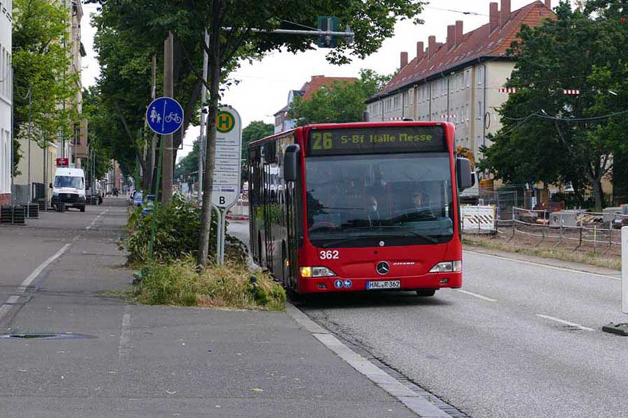 Roter Stadtbus der Linie 26 mit Zielanzeige 'S-Bf Halle Messe' fährt auf einer Straße neben einem Gehweg mit Fahrradweg-Schild und Haltestelle.