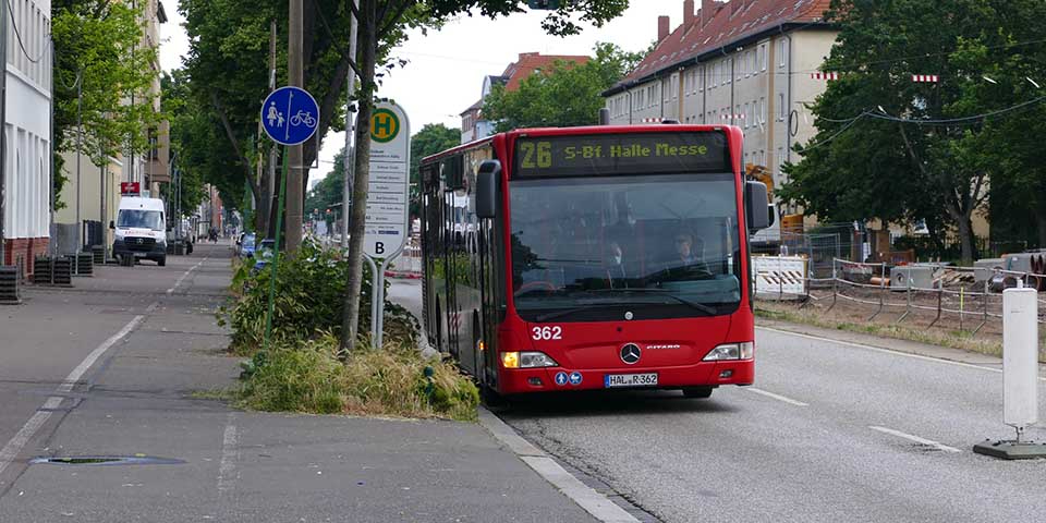 Roter Stadtbus der Linie 26 mit Zielanzeige 'S-Bf Halle Messe' fährt auf einer Straße neben einem Gehweg mit Fahrradweg-Schild und Haltestelle.