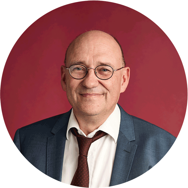 Mann mit Brille, weißem Hemd, dunkelroter Krawatte und dunkelblauen Anzug vor rotem Hintergrund