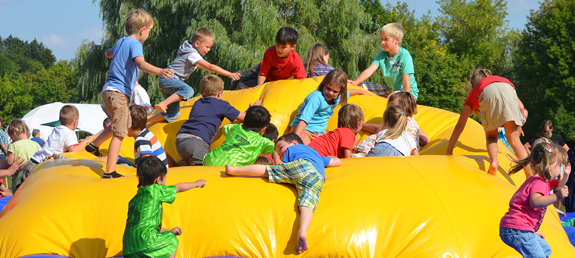 Auf dem Foto befindet sich mittig eine große gelbe Hüpfburg, auf der viele Kinder in bunter Kleidung spielen.