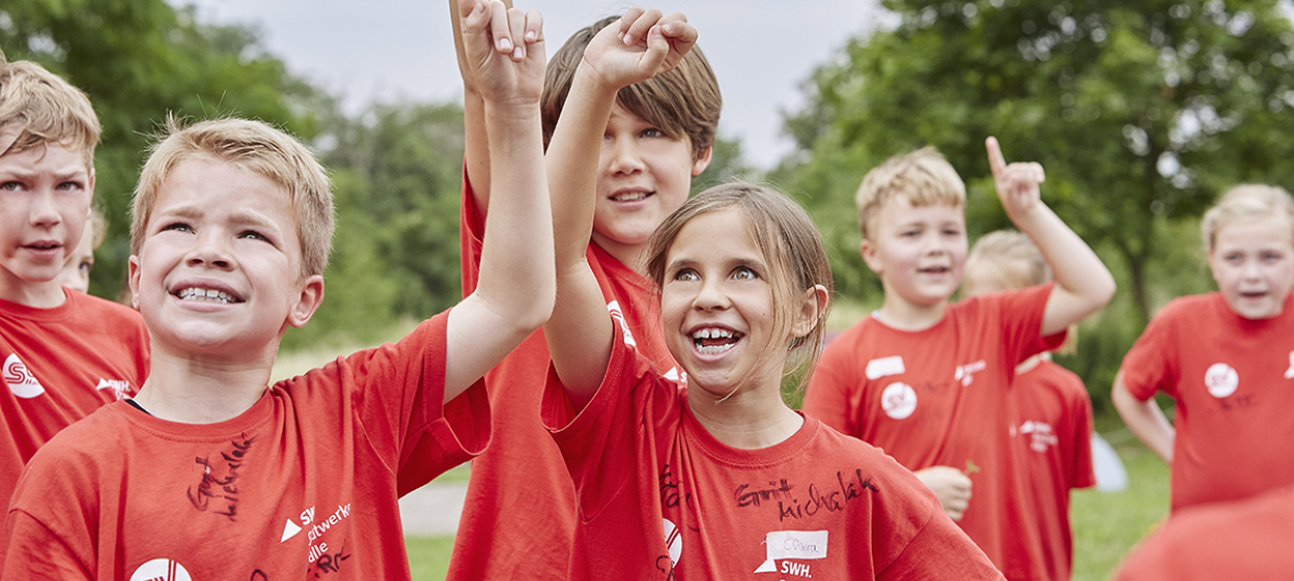 Das Bild zeigt eine Nahaufnahme von Kindern in roten T-Shirts, die auf einer Wiese mit Bäumen stehen und sich mit Fingerzeichen melden.