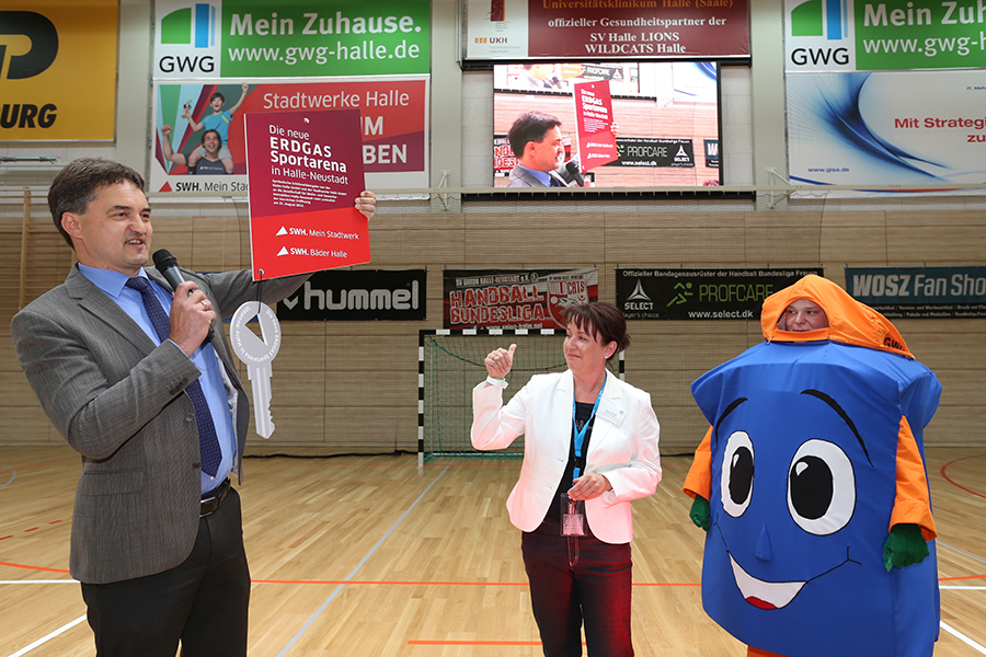 Feierliche Schlüsselübergabe in der Erdgas Sportarena durch Matthias Lux, Vorsitzender der Geschäftsführung der Stadtwerke Halle an Jana Kozyk, Geschäftsführerin der GWG. Rechts neben den beiden steht ein blaues Maskottchen.