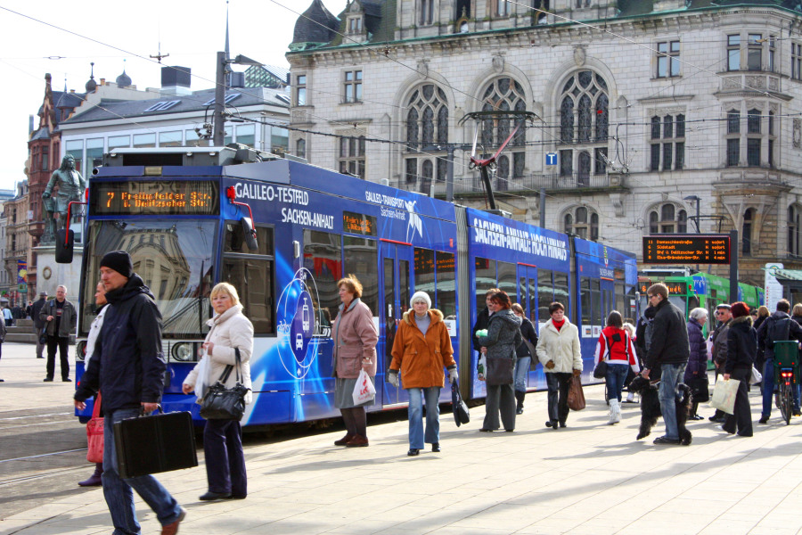 Auf dem Bild ist eine dunkelblaue Straßenbahn mit der Aufschrift "Galileo-Testfeld Sachsen-Anhalt" abgebildet. Sie steht an einer Haltestelle und um sie herum einige Menschen.