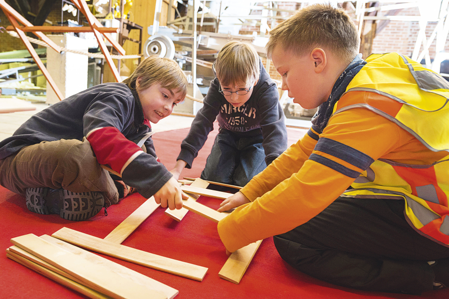 Hier sitzen drei Jungen auf einem roten Teppich und bauen eine Brücke aus Holzzuschnitten.