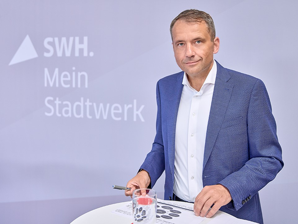 Mann im blauen Sakko steht an einem Tisch mit einem Blatt Papier und einem Stift vor einer Wand mit dem Schriftzug 'SWH. Mein Stadtwerk'.