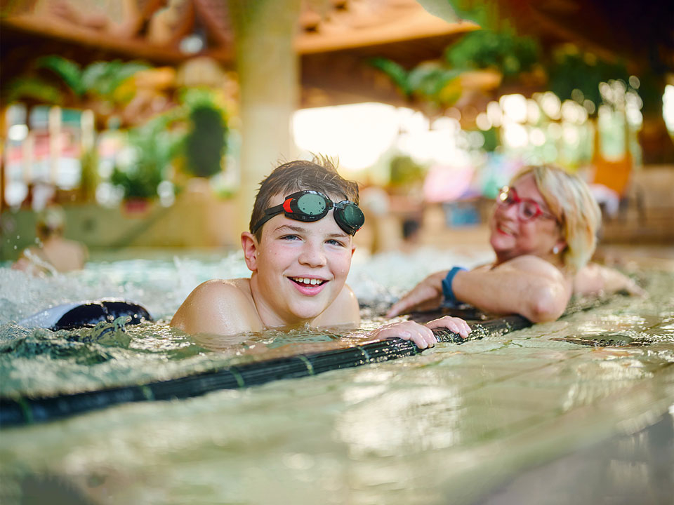 Junge mit Schwimmbrille hängt am Beckenrand und lächelt in die Kamera