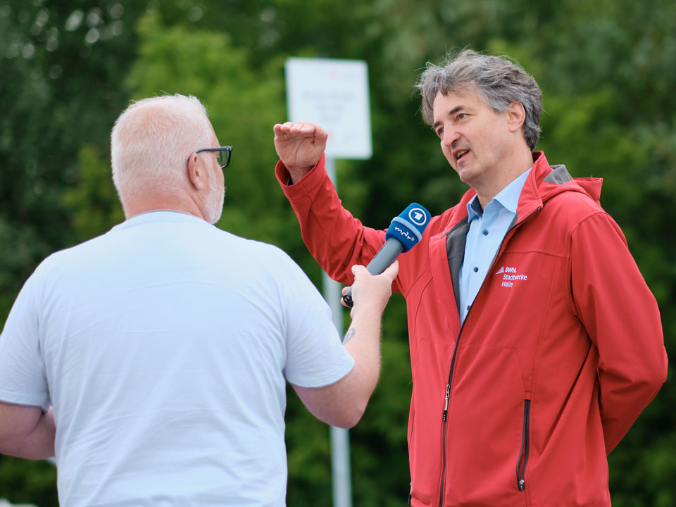 Zwei Männer im Freien. Einer mit weißem Haar und weißem T-Shirt hält ein Mikrofon, der andere trägt eine rote Jacke und gestikuliert mit der rechten Hand.