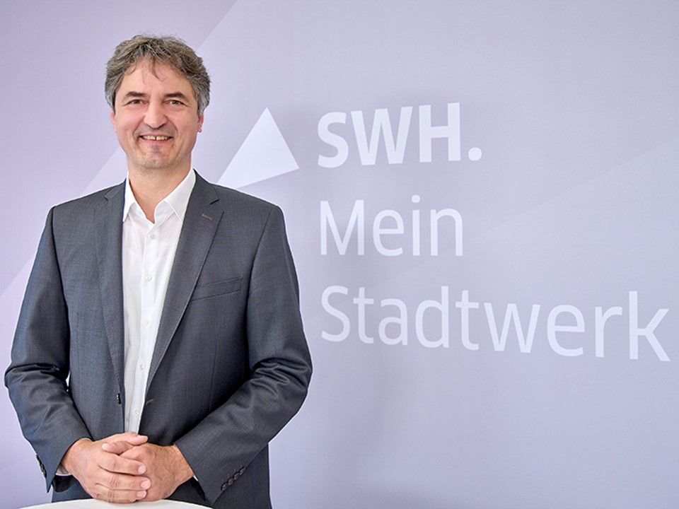 Mann im Anzug steht vor Wand mit Schriftzug 'SWH. Mein Stadtwerk' und lächelt