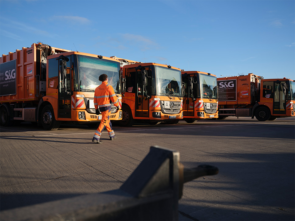 Person in orange-blauer Warnschutzkleidung steht vor einer Reihe orangefarbener Müllfahrzeuge.