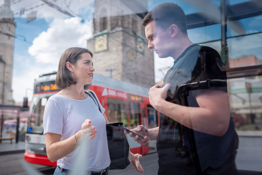 Zwei Personen stehen auf dem Marktplatz. Die Frau, welche links steht, trägt ein weißes T-Shirt und eine schwarze Handtasche über der Schulter. rechts steht ein junger Mann mit schwarzem T-Shirt. Beide unterhalten sich an einer Haltestelle. Im Hintergrund fährt eine rote Straßenbahn der HAVAG.