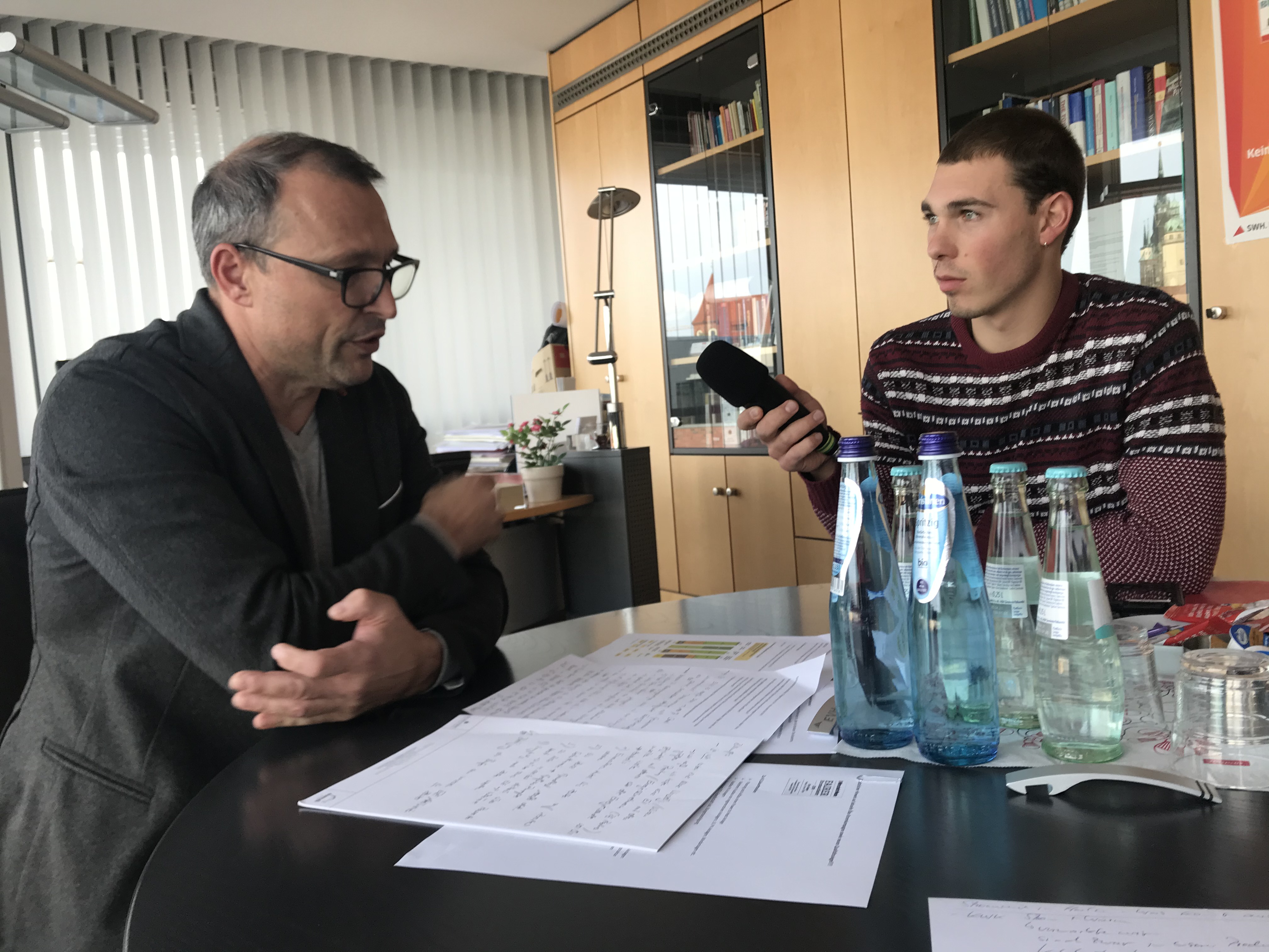 Das Bild wurde in einem Büro aufgenommen. Vorne befindet sich ein Tisch mit Wasserflaschen und Papieren und im Hintergrund eine Schrankwand und ein Fenster mit Jalousien. Auf der linken Seite sitzt Olaf Schneider, ein Herr mit grauem Anzug und Brille, und rechts Maximilian Hitzler, der einen rotgestreiften Pulli trägt und ein Mikrofon in Richtung seines Gesprächspartners hält.