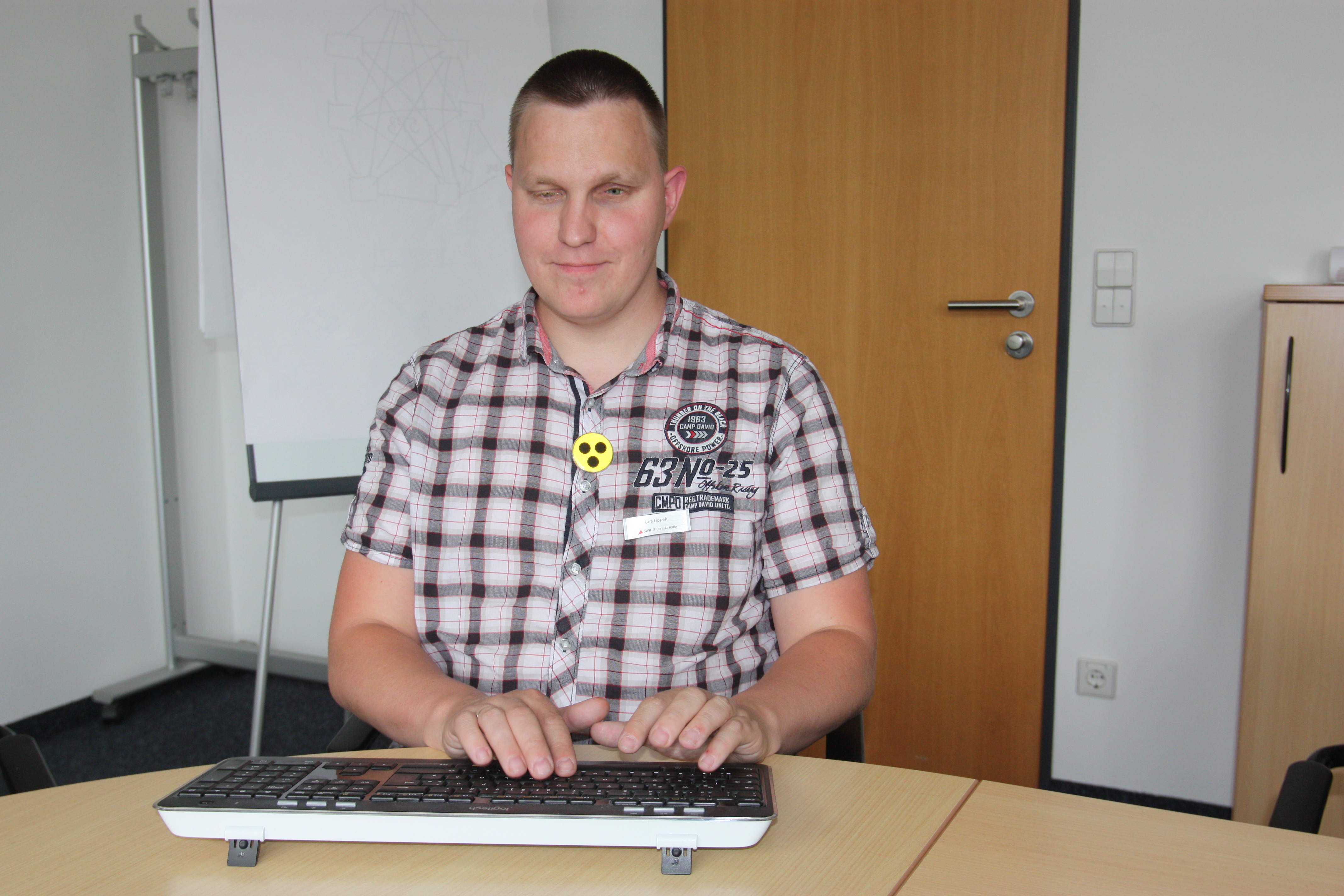 Das Bild zeigt den blinden Lars Lippek sitzend am Tisch. Vor ihm steht seine mit einer Braillezeile ausgestattete Tastatur. Im Hintergrund ist ein normales Büro zu sehen.