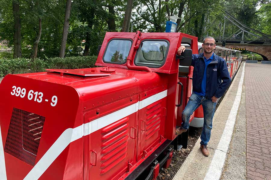 Das Foto zeigt mittig den Peißnitzexpress und rechts davon einen Mann in blauer Kleidung, der sich an die Lok der Parkeisenbahn anlehnt.