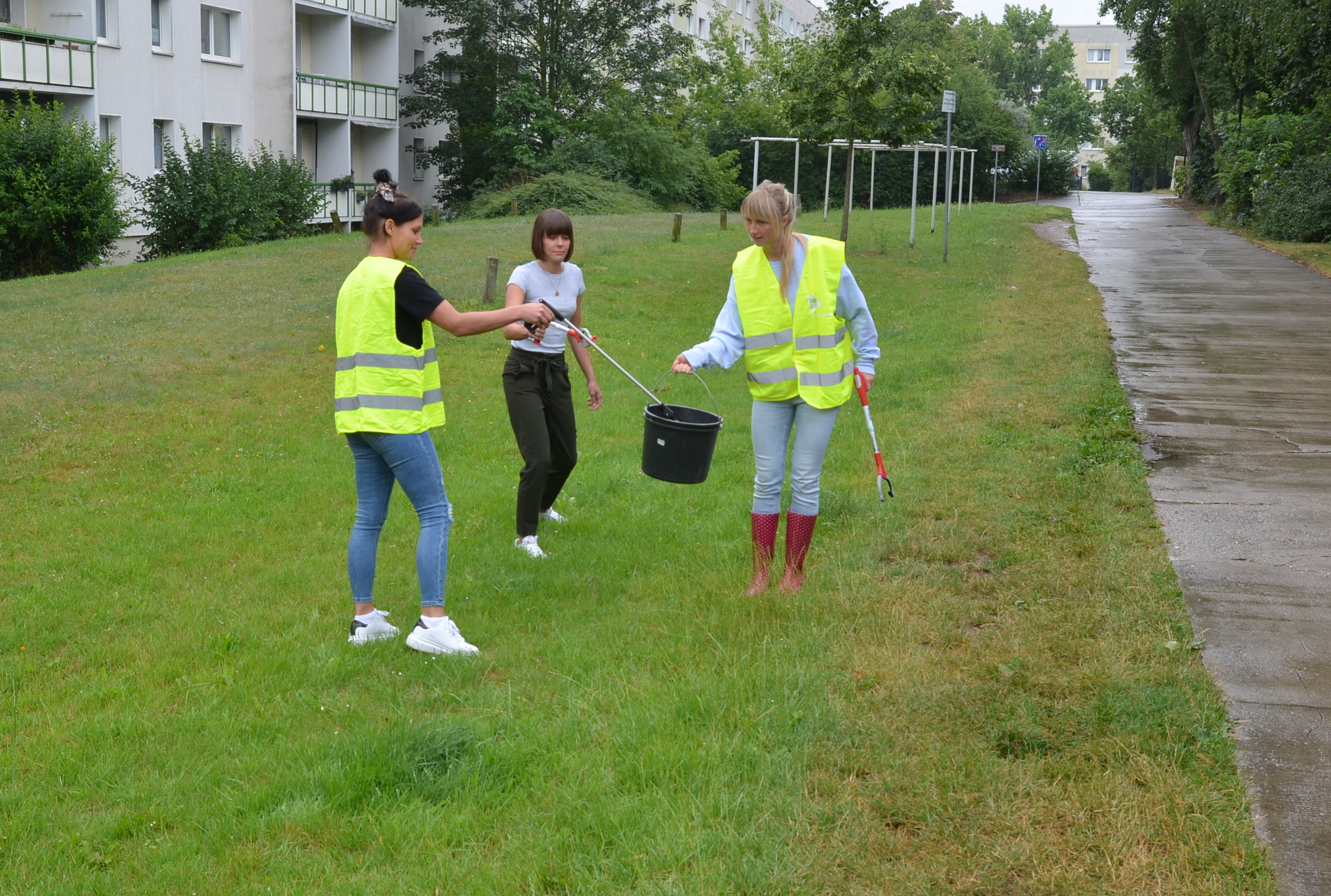 Das Foto zeigt drei Frauen, von denen zwei eine gelbe Warnweste tragen. Sie alle halten Müllgreifer in den Händen und sammeln Müll auf einer grünen Wiese.
