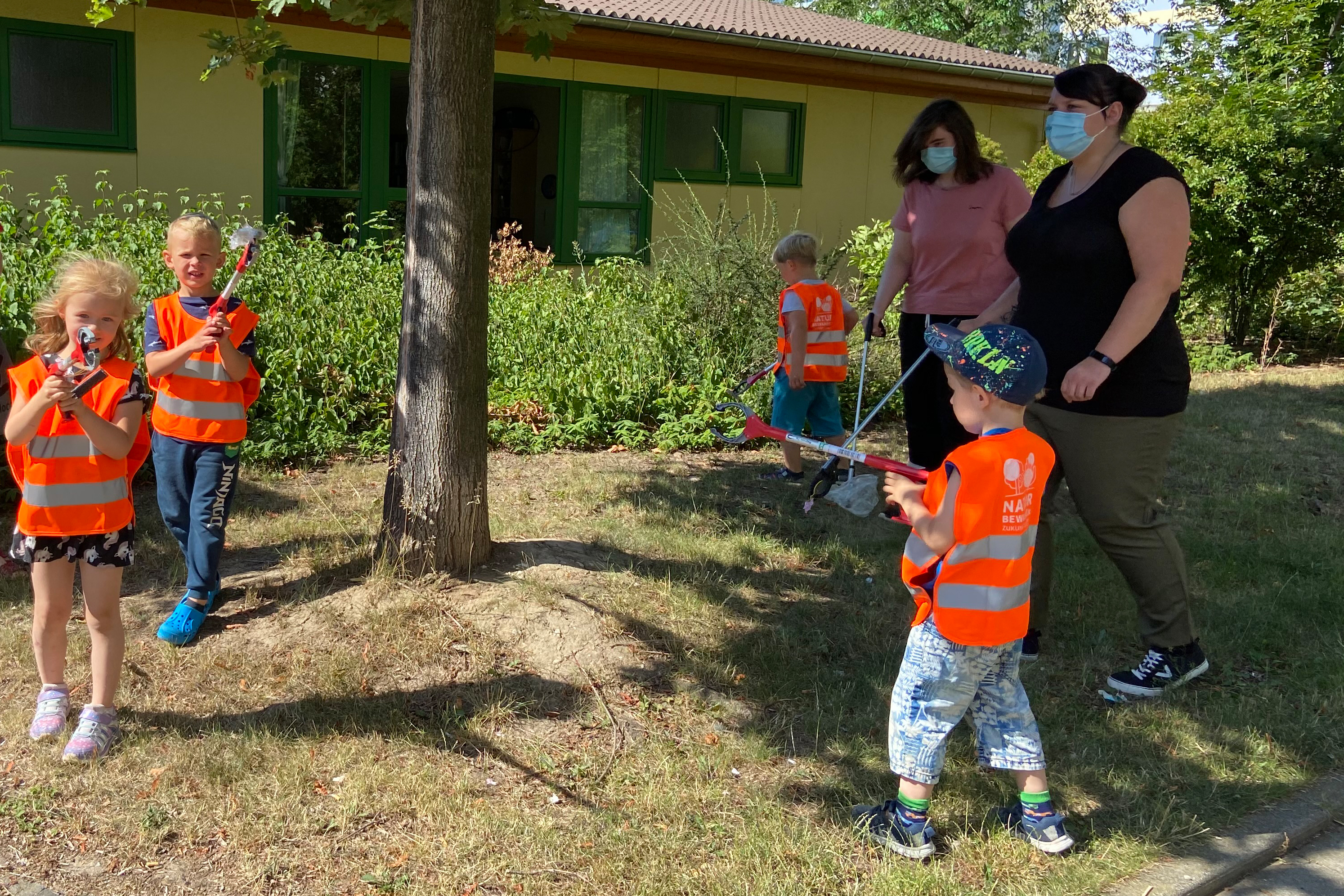 Zwei Erzieherinnen und vier Kinder laufen mit Müllgreifern in den Händen über eine Wiese auf der Suche nach Müll. Im Hintergrund steht ein gelbes Haus.