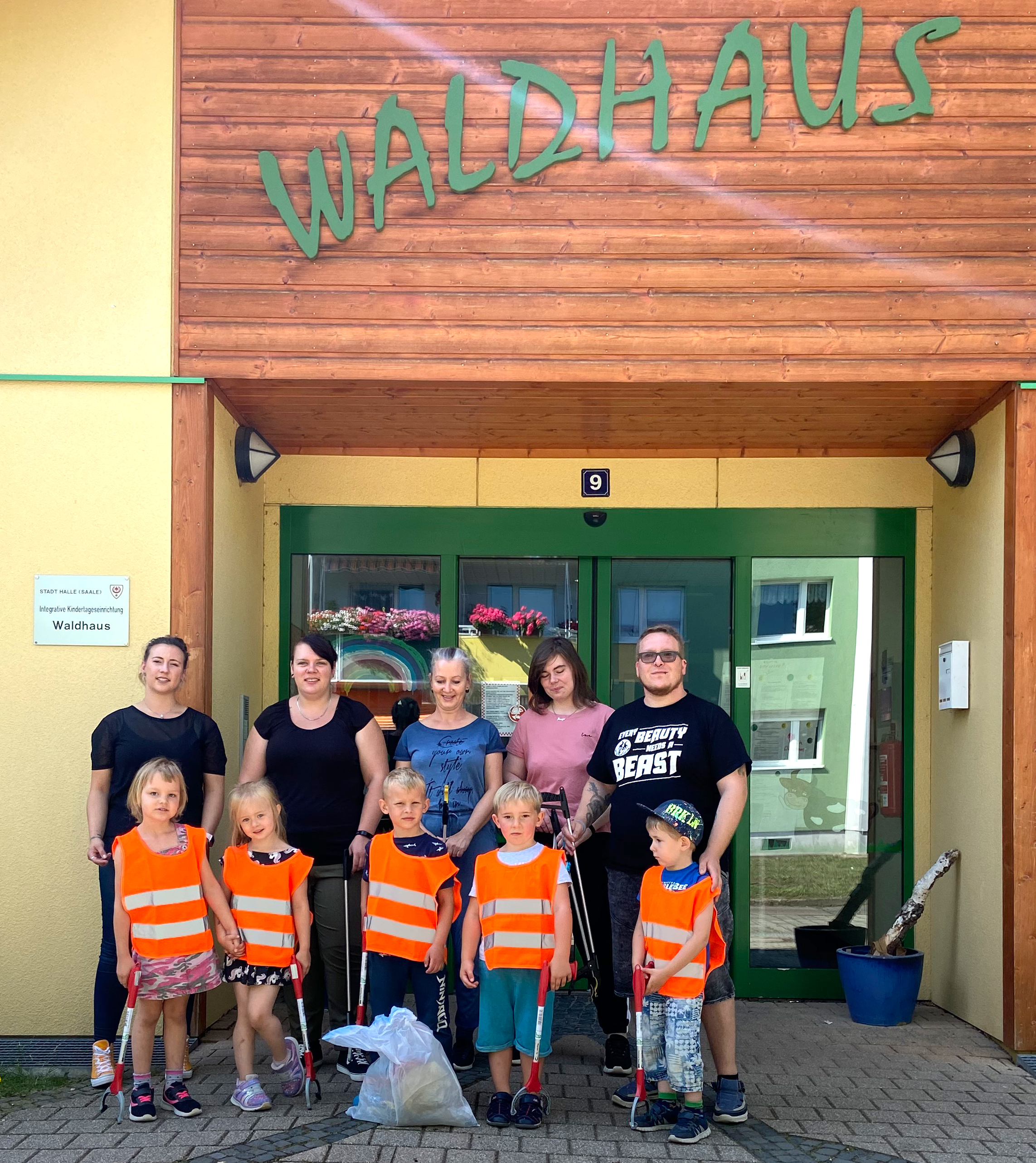 Das Bild zeigt fünf Erwachsene und fünf Kinder mit Warnweste und Müllgreifern in den Händen vor einer Eingangstür, über der in grünen Buchstaben "Waldhaus" steht. Vor den Kindern steht ein weißer Müllsack.