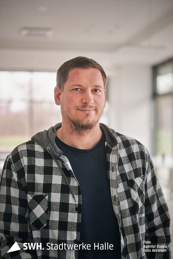 Eine Portraitaufnahme eines jungen Herren. Er lächelt in die Kamera trägt ein dunkelblaues T-Shirt und darüber ein kariertes Hemd.