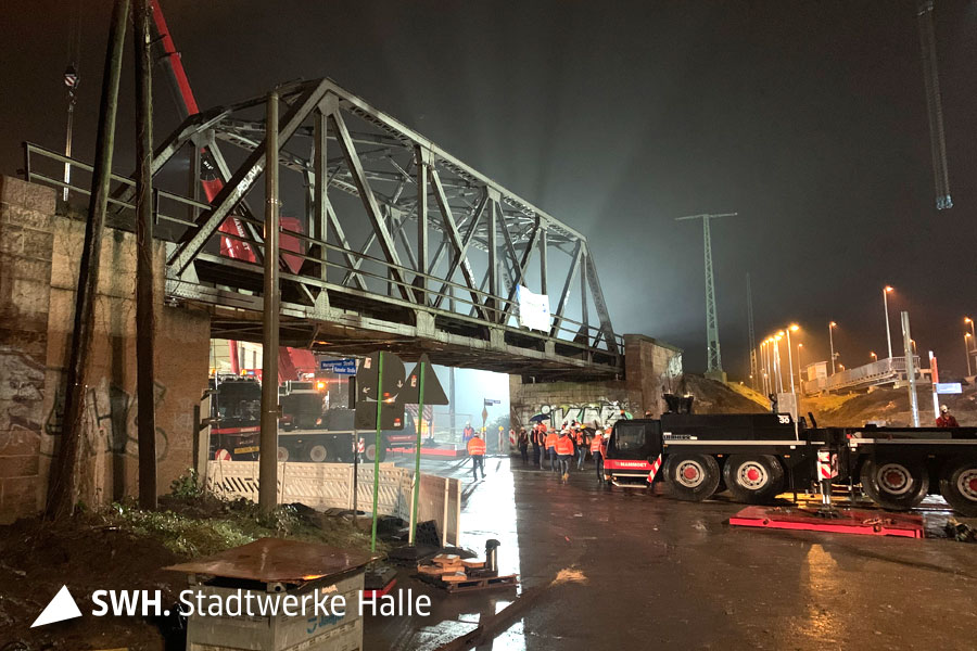 Eine Nachtaufnahme der Baustelle. Die alte Rosengartenbrücke wird angestrahlt. Einzelne Mitarbeiter im leuchtend orangefarbenen Jacken stehen an der Baustelle. Im Hintergrund ist ein Kran zu sehen. Im Vordergrund ein großer Laster. 