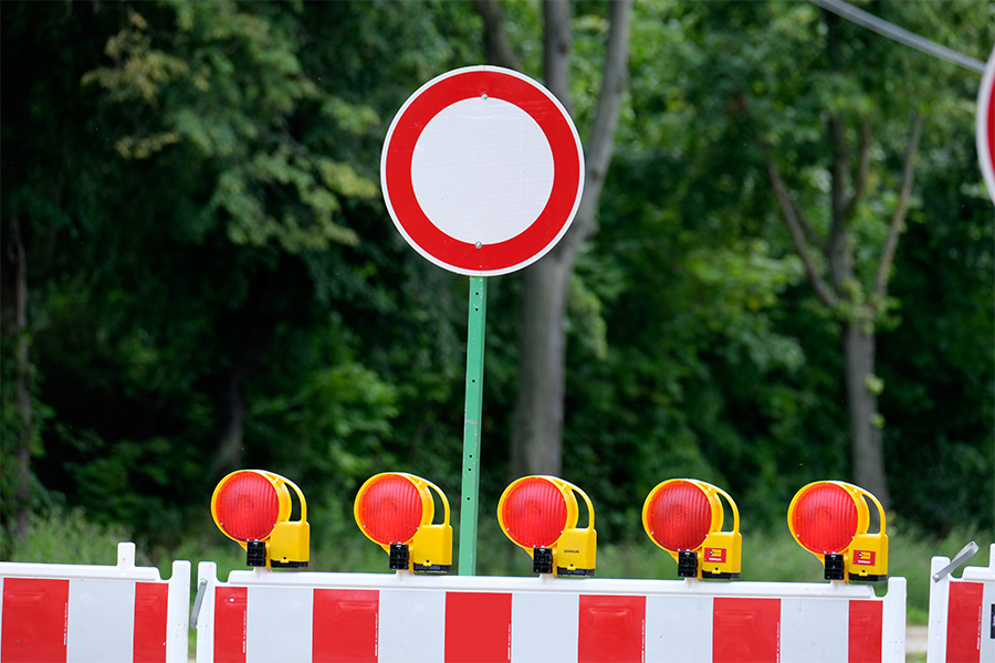 Das Bild zeigt ein "Durchfahrt verboten"-Verkehrsschild. Im Hintergrund sind Bäume unscharf zu sehen. Im Vordergrund eine Baustellenabsperrung mit roten Lichtern.