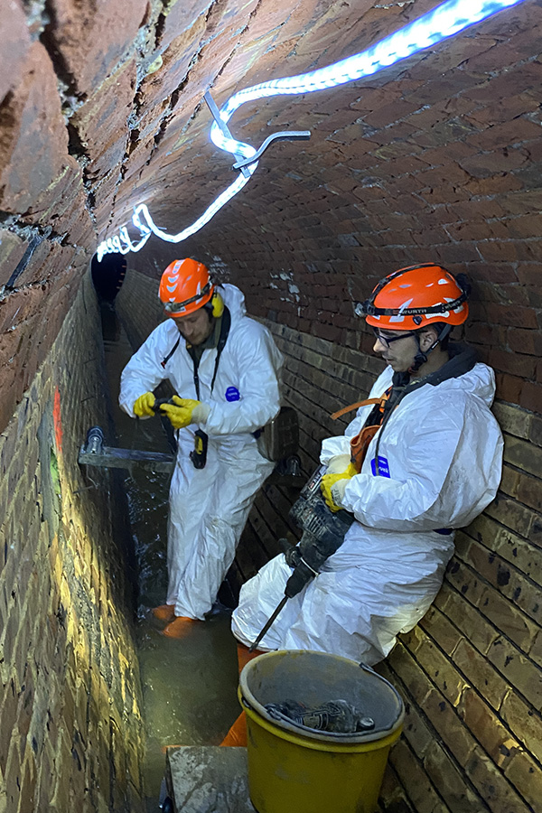 Das Bild zeigt zwei Männer in weißen Anzügen, die mit Hilfe von Geräten die Mauer des Kanals untersucht.