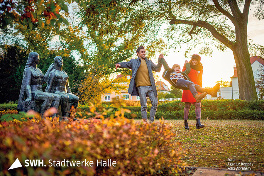 Eine Familie, bestehend aus Vater, Mutter und Kind, haben Spaß im Grünen. Am linken Bildrand ist eine Statue zu sehen. Im Hintergrund ein Haus.