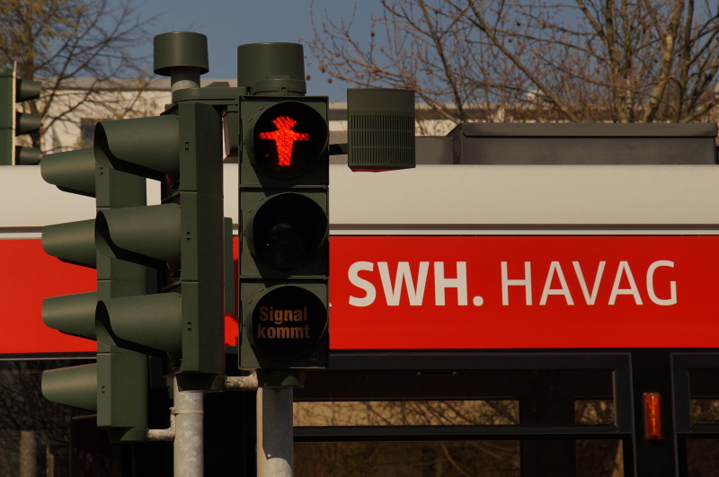 Auf dem Bild befindet sich mittig eine Fußgängerampel, die ein rotes Männchen zeigt. Rechts daneben ist der Tonsignalgeber angebracht. Im Hintergrund steht ein roter Bus mit der weißen Aufschrift SWH.HAVAG.