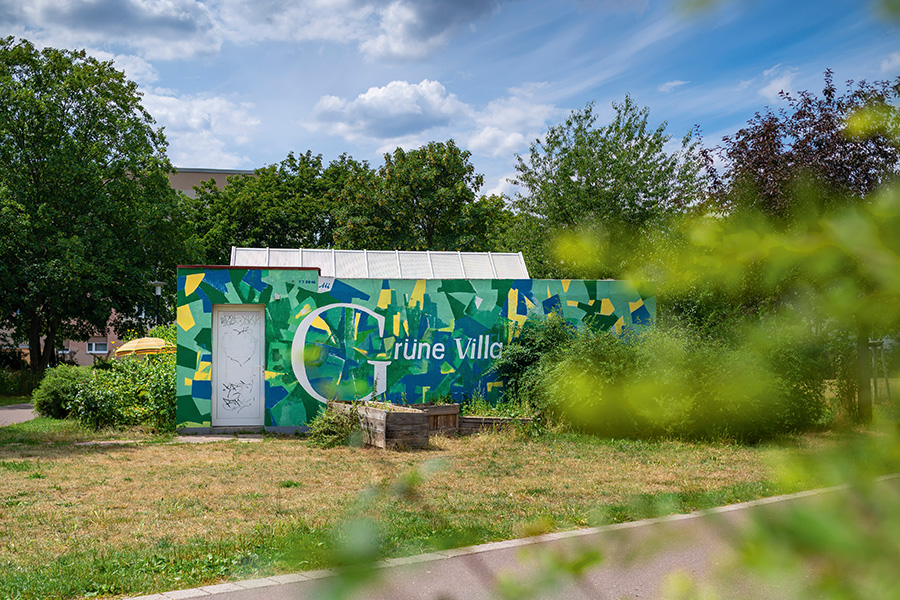 Das Bild wurde in einem Garten aufgenommen und zeigt ein kleines flaches Gebäude, das grün, gelb und blau angemalt wurde und mit dem großen weißen Schriftzug "Grüne Villa" verziert ist.
