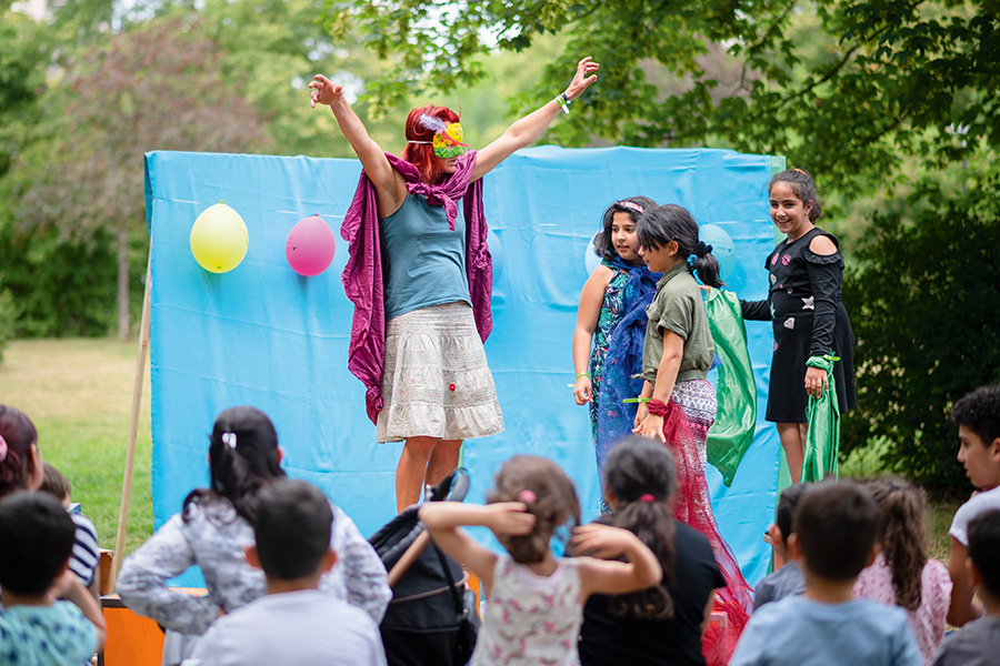 Im Bildmittelpunkt stehen eine Frau und drei Mädchen. Sie stehen auf einer Bühne vor einer türkisen Kulisse und sind verkleidet. Im Vordergrund sitzen mehrere Kinder, die die vier Personen auf der Bühne beobachten.