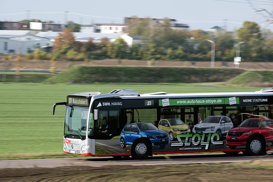 Ein silberner Bus der HAVAG fährt auf der Straße von links nach rechts durch das Bild. Im Vordergrund ist Acker zu sehen, im Hintergrund Wiese, Bäume und eine Straße. Der Fokus liegt auf dem Bus, der Hintergrund ist unscharf. 