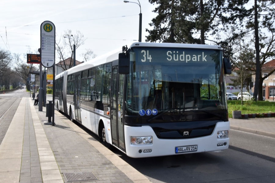 Ein weißer Bus an einer Haltestelle. Der Himmel ist bedeckt. Im Hintergrund sind Nadelbäume.