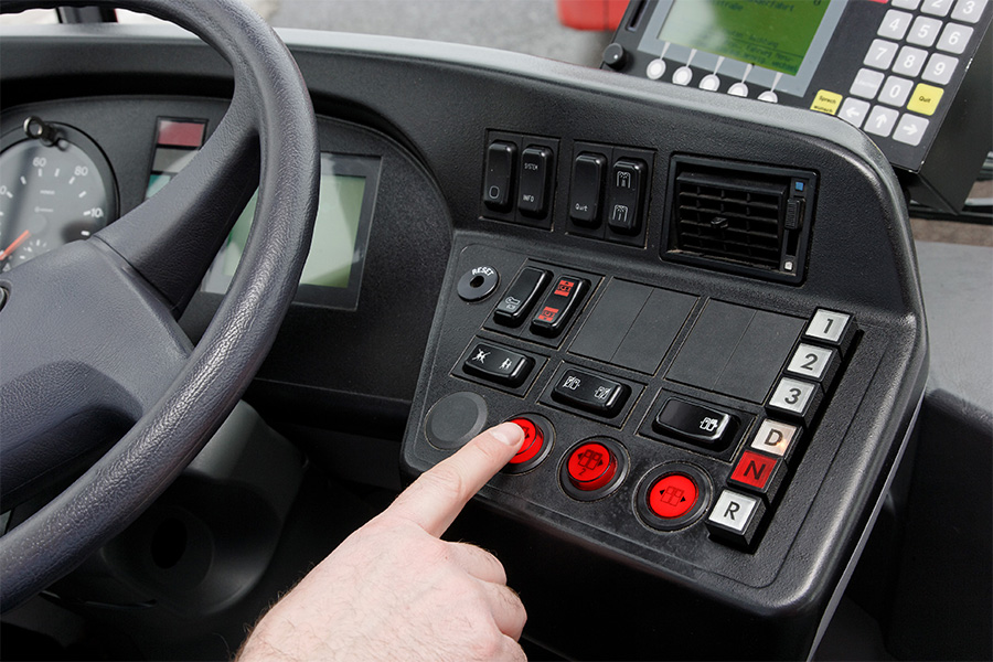 Ein Finger drückt einen Knopf am Bedienpult neben dem Lenkrad in einem Bus.