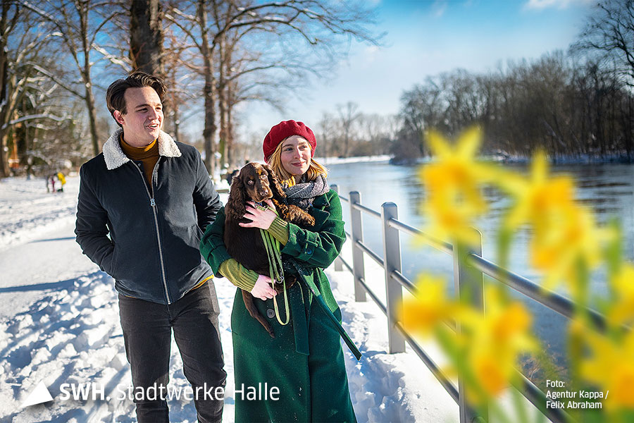 Eine Mann und eine Frau gehen an der Saale spazieren. Schnee liegt, die Sonne scheint. Die Frau trägt einen grünen Mantel, eine rote Mütze und einen Hund auf dem Arm. Der Mann läuft neben ihr.