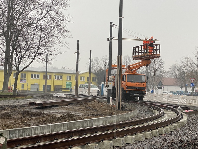 Auf diesem Foto sind mittig neue Straßenbahn-Gleise abgebildet. Im Hintergrund befindet sich ein großes orangefarbenes Hubfahrzeug, auf dem zwei Personen stehen, die an den Oberleitungen arbeiten.