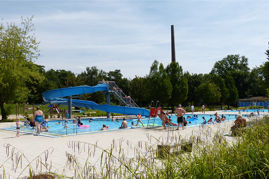 Das Freibad Saline im Sommer bei blauem Himmel. Einige Personen stehen am Beckenrand, einige sind im Wasser, machen an der Rutsche. Die Rutsche ist blau. Im Vordergrund sind Grashalme zu sehen.