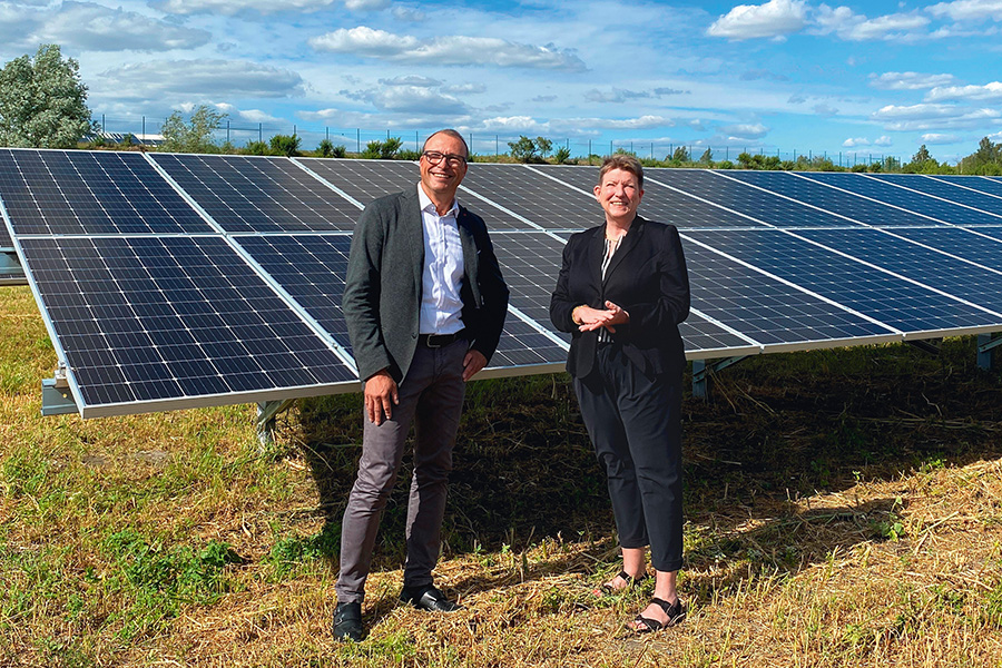 Im Bild stehen ein Mann in grauem Anzug und Brille und eine Frau in schwarzem Anzug vor einem Solarfeld und lächeln in die Kamera.