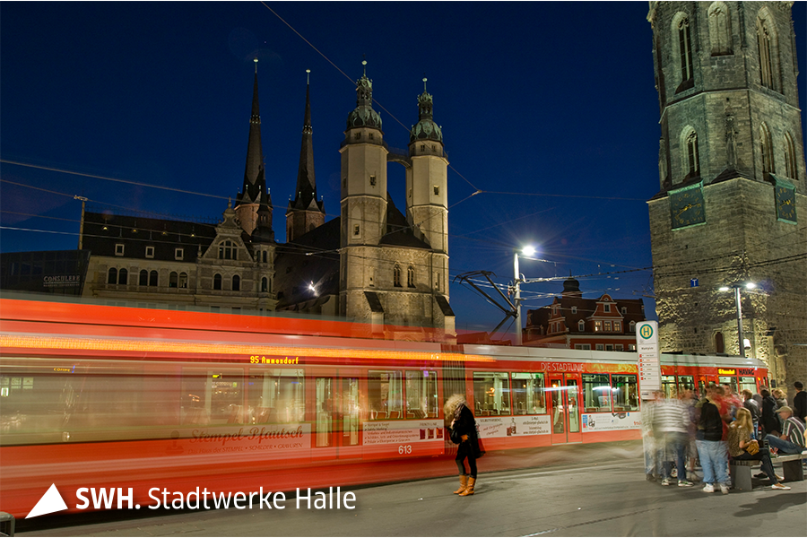 Eine Straßenbahn abends am zentralen Marktplatz in Halle (Saale). Im Hintergrund sind die ikonischen Türme der Stadt zu sehen. Durch die Langzeitbelichtung sind sowohl die Straßenbahn als auch die Menschen verschwommen zu erkennen.