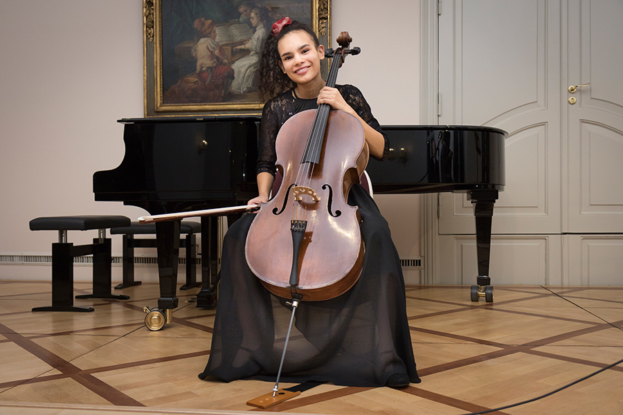 Auf dem Bild ist die Gewinnerin der Händel-Mozart-Jugendstipendien Sophia Hartmann zu sehen. Sie sitzt auf einem Stuhl und hat ihr Cello aufrecht vor sich gestellt. Ihre braunen, lockigen Haare trägt sie in einem Zopf. Sie trägt ein schwarzes Kleid. Im Hintergrund ist ein Gemälde zu sehen. 