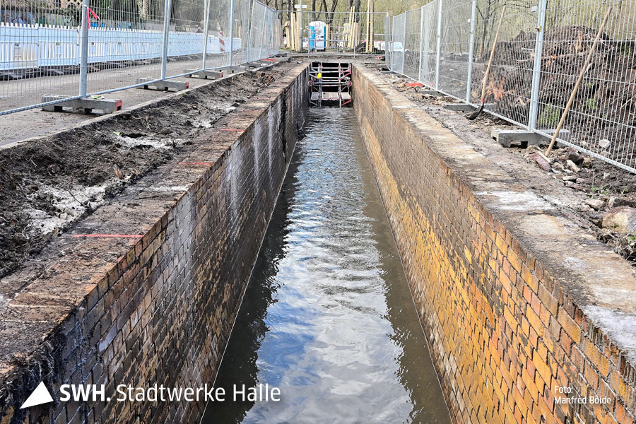 Blick in einen etwa ein Meter breiten gemauerter Kanal mit Wasser. Die Mauersteine sind gelb und dreckig. Der Abwasserkanal hat keine Betondecke mehr. Man sieht das Wasser. Links und rechts davon steht ein Bauzaun.