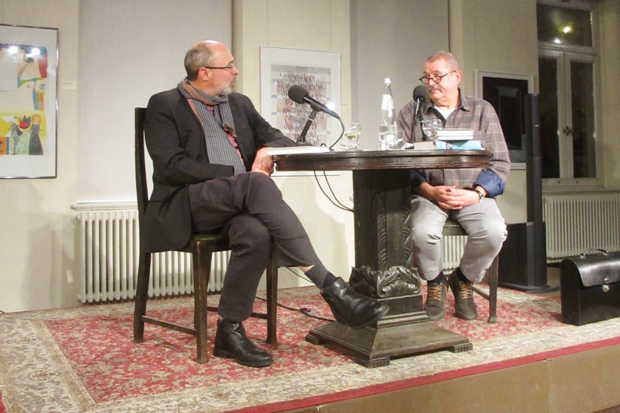 Das Bild zeigt zwei Männer, die an einem Tisch sitzen, auf dem ein Mikrofon steht.