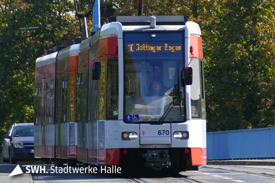 Die rote Straßenbahn der HAVAG fährt direkt auf den Fotografen zu. Die Bahn ist frontal zu sehen und kommt aus einer leichten Rechtskurse. Hinter der Bahn ist ein dunkles Auto. Im Hintergrund sind grüne Bäume mit Laub. Rechts der Bahn ist ein hellblaues Geländer.