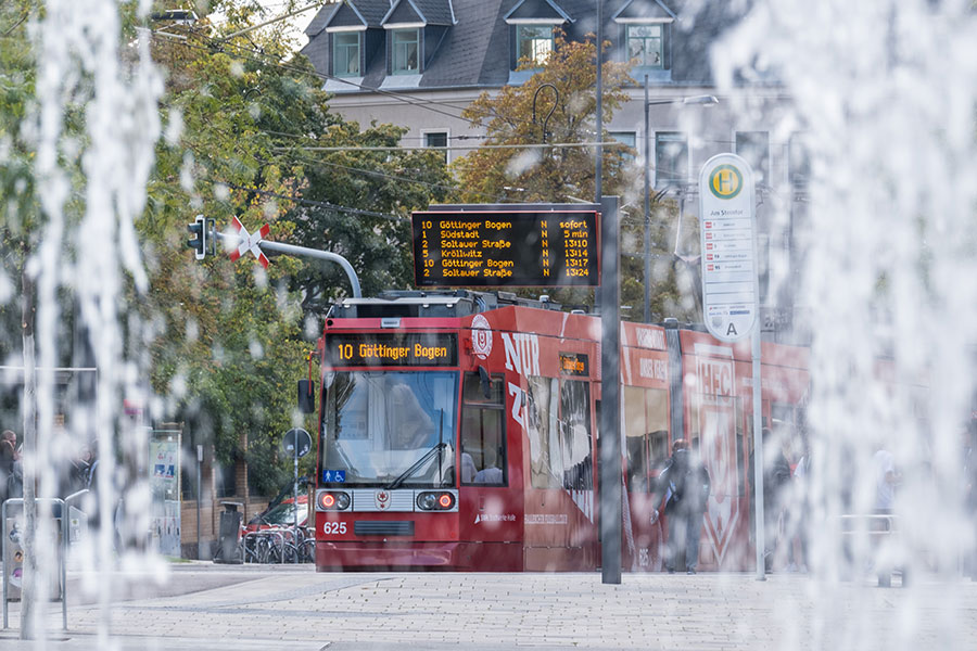 Die rote Straßenbahn der Linie 10, mit der Zielaufschrift "10 - Göttinger Bogen" fährt seitlich auf den Fotografen zu. Der Fotograf fotografiert druch einen Springbrunnen hindurch.