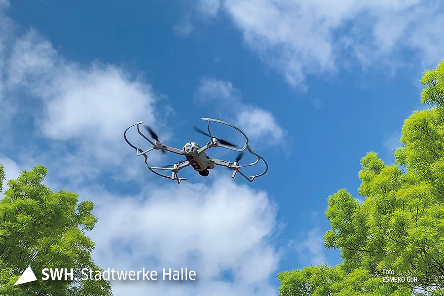 Eine Drohne wird von unten fotografiert. Im Hintergrund ist strahlend blauer Himmel mit ein paar weißen Wolken zu sehen. Außerdem sind Bäume zu sehen. Das Bild ist aus der Froschperspektive aufgenommen.