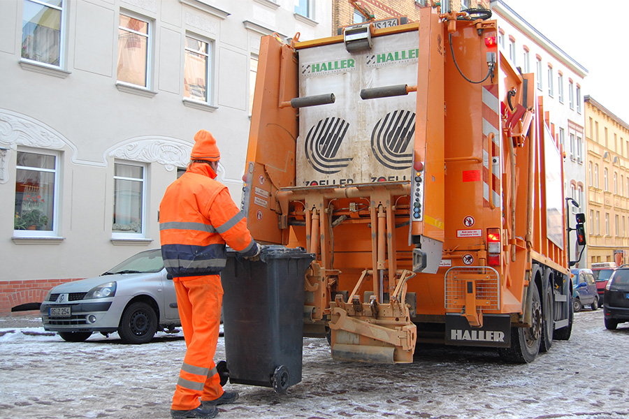 Ein Morangefarbenes Müllauto und ein Mitarbeiter der HWS mit orangefarbener Kleidung sind von hinten zu sehen. Der Mann trägt eine Orangefarbene Mütze und mit seinen Händen schiebt er eine Mülltonne an das Auto. Sie stehen n einer leicht verschneiten Straße. Im Hintergrund ist ein weißes Haus mit mindestens 3 Etagen.