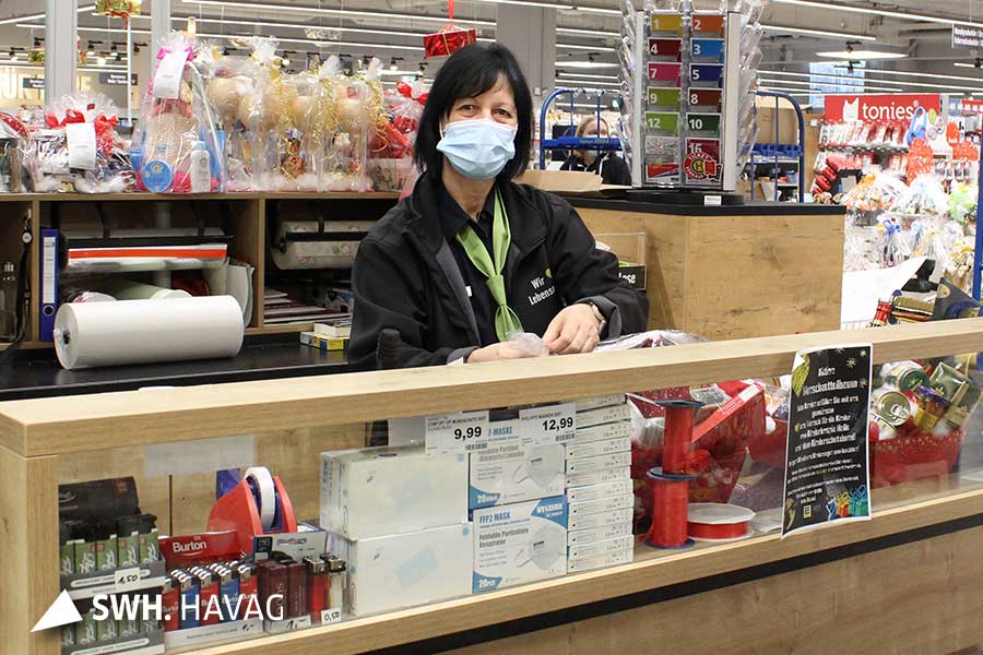Eine Frau mit dunklen Haaren steht in Arbeitsbekleidung an einem Tresen. Im Hintergrund ist der Laden zu sehen. Die Dame blickt in die Kamera.