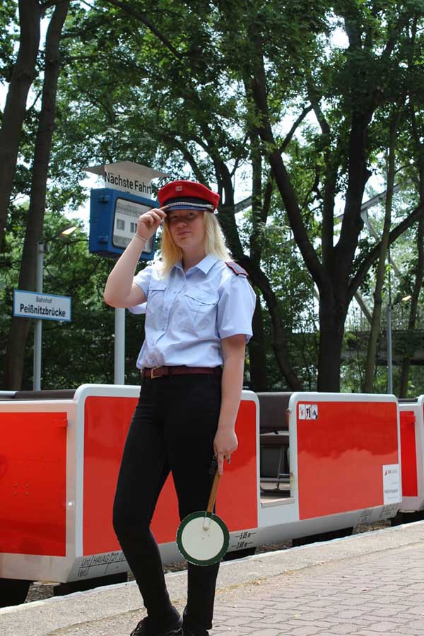 Auf diesem Foto steht ein blondes Mädchen mit Lokführer-Uniform und Kelle in der Hand vor den roten Waggons des Peißnitzexpress.