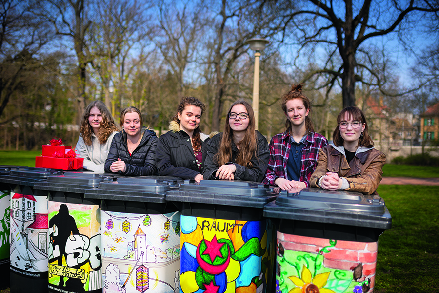 Sechs junge Damen stehen hinter den neu gestalteten Mülltonnen. Alle schauen in die Kamera. Das Bild wurde auf der Peißnitz aufgenommen. Im Hintergrund ist grüner Rasen und Bäume zu finden. 