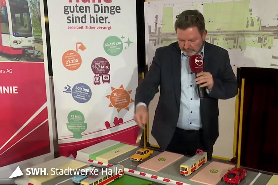 Erhard Krüger trägt einen dunklen Anzug und hält ein rotes Mikro von TV Halle in seiner linken Hand. In seiner rechten Handy zeigt er auf den Tisch vor ihm. Er blickt nach unten. Auf dem Tisch stehen Autos und eine Straßenbahn sowie Bausteine, die die neuen Aufteilung der Straße symbolisieren.