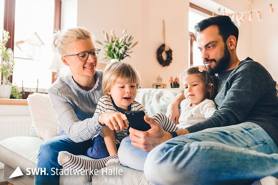 Ein Mann sitzt mit seiner Frau und seinen 2 kleinen Kindern auf der Couch und schaut sich gemeinsam das Handy an.