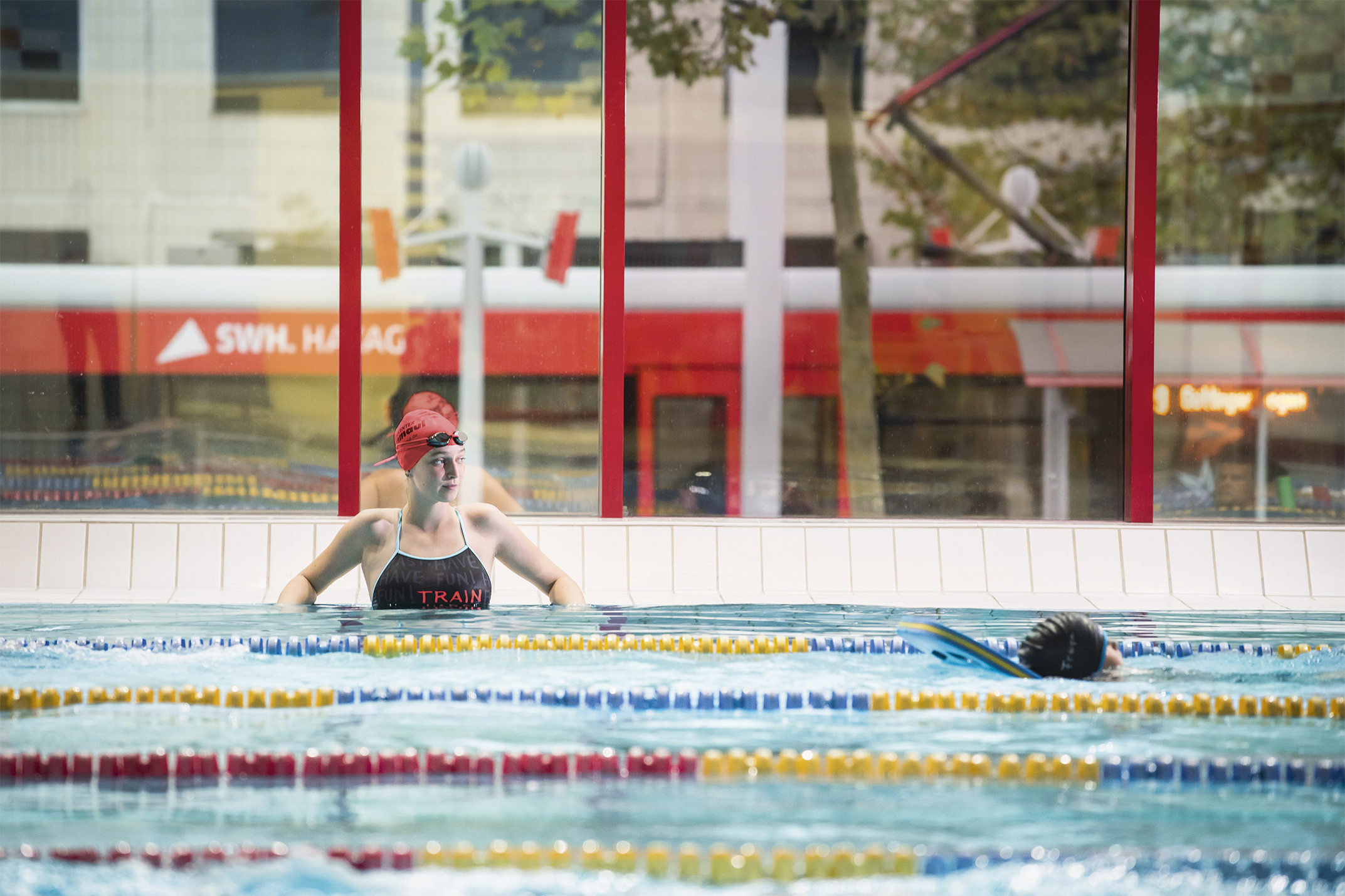Das Bild zeigt eine Schwimmerin im Wasser in der Schwimmhalle Neustadt. Durch die großen Fenster im Hintergrund ist die Straßenbahn zu sehen.